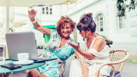 Two happy senior women using laptop and pointing at screen surprised while sitting in outdoor cafeの写真素材