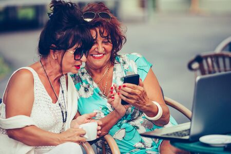 Two happy senior women friends are using a smart phone and talking while sitting outdoors in a cafeの写真素材