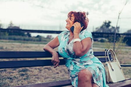 Senior beautiful woman is calling on a cell phone and talking while sitting on a benchの写真素材