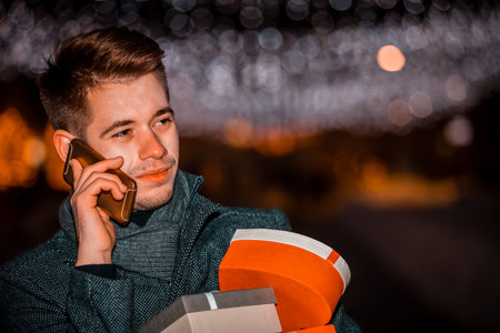 Handsome man talking on the mobile phone and holding presents at night in decorated city street.