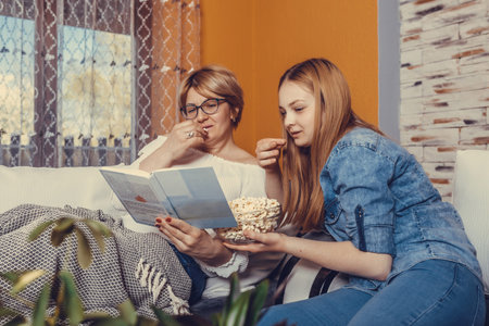 Mother and her teenage daughter are reading book and eating popcorn while sitting on the couch at...