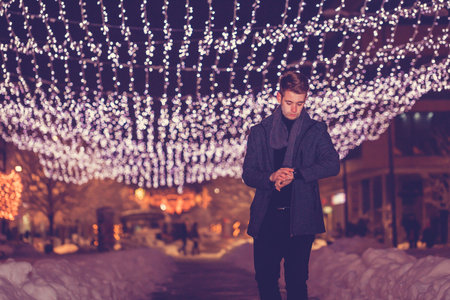 Handsome young man looking at his watch and waiting for someone while standing  at night in...