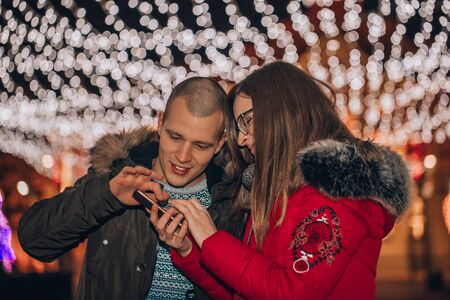 Cute couple watching at smartphone screen together, against the backdrop of city lights.の写真素材