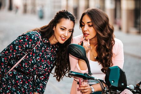 Two young beautiful women putting lipstick looking in mirror, while sitting on the scooter.の写真素材