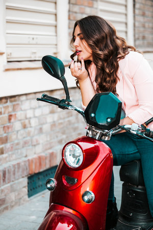 Young beautiful woman putting lipstick looking in mirror, while sitting on her scooter.