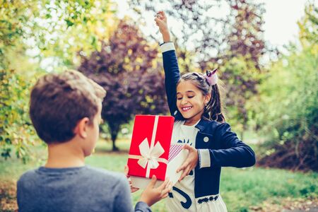 Happy little child boy gives smiling girl a gift box, making a surprise. Happy little girl rejoices gift she received from her best friendの写真素材