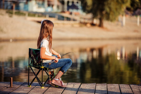 Little Caucasian girl sitting on a wooden pier with fishing rod. Cute child fishing on a lake in a sunny summer day.の写真素材
