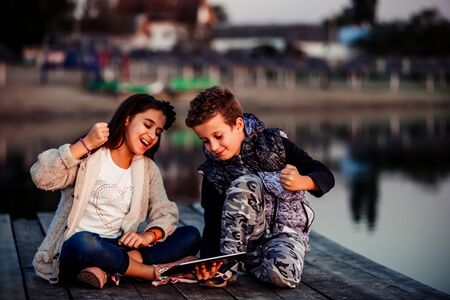 Two young cute little friends, boy and girl having fun playing on digital tablet sitting by the lake in the evening. Children celebrating success. Friendshipの写真素材