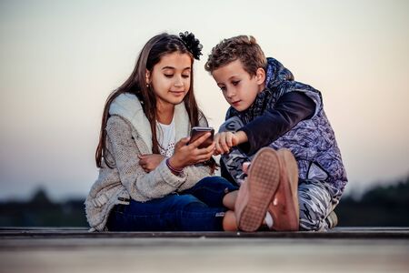 Two young cute little friends, boy and girl watching something on smartphone sitting by the lake in the evening. Children having fun together. Friendshipの写真素材
