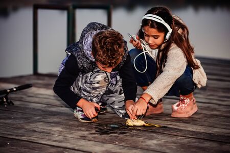 Two young cute little friends, boy and girl fishing on a lake in the evening. Little boy showing his friend a fish caught. Kids are playing. Friendship.の写真素材