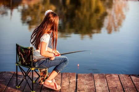 Little Caucasian girl sitting on a wooden pier with fishing rod. Cute child fishing on a lake in a sunny summer day.の写真素材