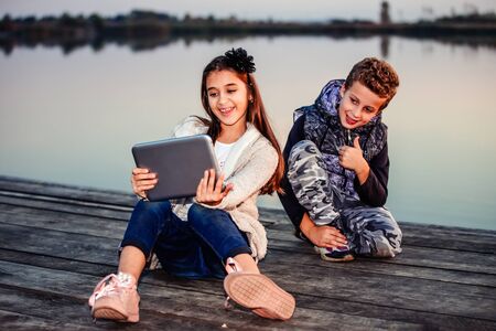 Two young cute little friends, boy and girl having fun and  making selfie for blogging on digital tablet while sitting by the lake in the evening. The boy showing gesture thumb up. Friendshipの写真素材