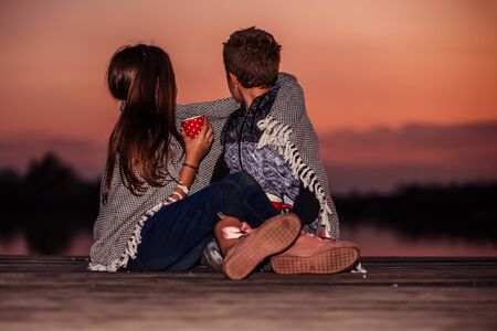 Two young cute little friends, boy and girl talking while sitting covered with blanket by the lake during sunset.の写真素材