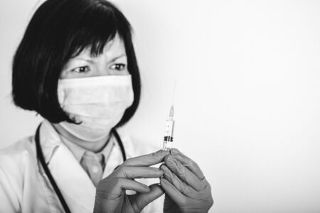 Female doctor in white lab coat and medical mask holding a syringe with medicine in his hand, an injection. Black and white photo.の写真素材