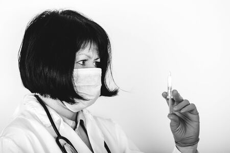 Female doctor in white lab coat and medical mask on white background holding syringe and medicine in ampoule. Black and white photo.の写真素材