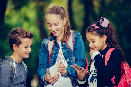 Outdoor portrait of positive girls and boy playing with phones after school while walking through the park.の写真素材