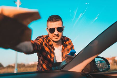 Young man carefully cleaning a windshield on his car with microfiber cloth and spray in a bottle.の写真素材
