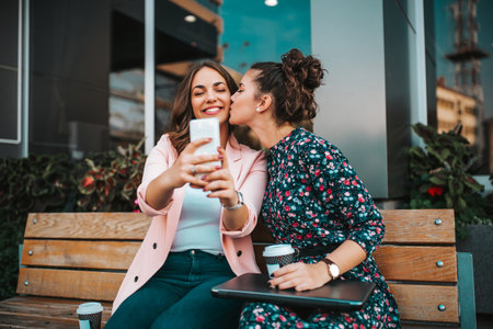Two joyful cheerful girls taking a selfie while sitting together in the city street. Happy girl...