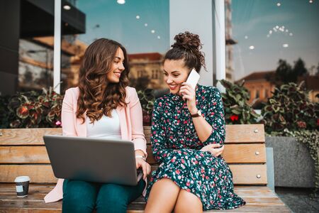 Two cheerful female friends searching media content online in a laptop and talking on mobile phone while sitting on the bench in the city streetの写真素材