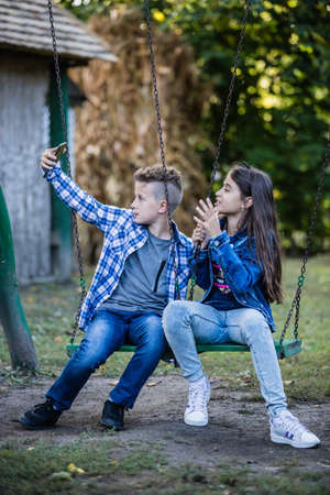 Little cute boy and girl are taking picture of themselves for selfie with phone while sitting on swingの写真素材