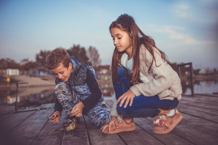 Two young cute little friends, boy and girl fishing on a lake in the evening. Little boy showing his friend a fish caught. Kids are playing. Friendship. Selective focus.の写真素材