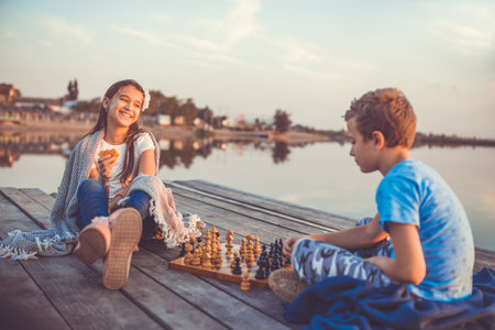 Two young cute little friends, boy and girl having fun while playing chess sitting by the lake in the evening.の写真素材