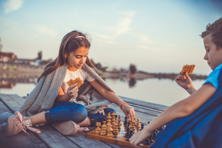 Two  little friends, boy and girl having fun while playing chess sitting by the lake in the evening. Kids are playing. Friendship. Selective focus.の写真素材