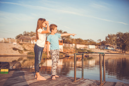 Two young cute little friends, boy and girl pointing with finger away on a lake in a sunny summer day. Kids are playing. Friendship.  Selective focus.の写真素材
