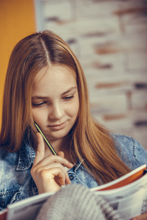 Cute teenage girl sitting on a couch and studying before the exam, she is reading a book and writing...