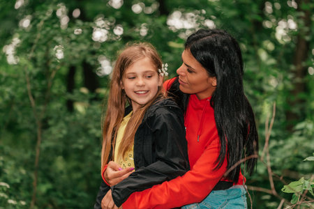 Beautiful mother and her cute daughter sitting in forest and talking, spending good time in spring nature. Mother is hugging her daughter.の写真素材