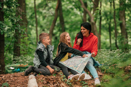 Beautiful mother with two kids are having break in the forest, using a map and planning a hiking adventure. Happy family having fun together. Selective focus.の写真素材