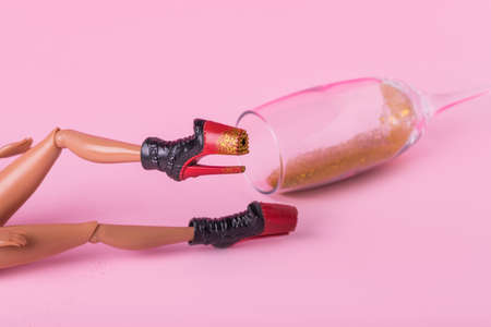 Minimal composition with female doll laying on the floor with trendy high heel and spilled champagne glass full of gold glitter on pink background. Creative minimal after party concept.の写真素材