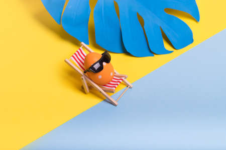 Creative funny composition with Easter egg with sunglasses while sitting on deck chair on illuminating yellow and pastel blue background. Trendy minimal  spring or summer vacation, holiday and travel concept.の写真素材