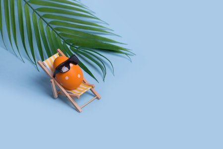 Creative funny composition with Easter egg with sunglasses sitting on deck chair and palm leaf on...