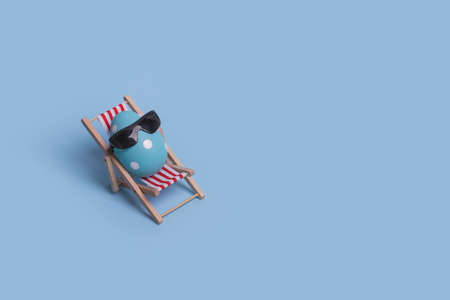 Creative funny composition with Easter egg with sunglasses while sitting on deck chair on bright blue background. Trendy minimal  spring or summer vacation, holiday and travel concept. Free space for textの写真素材