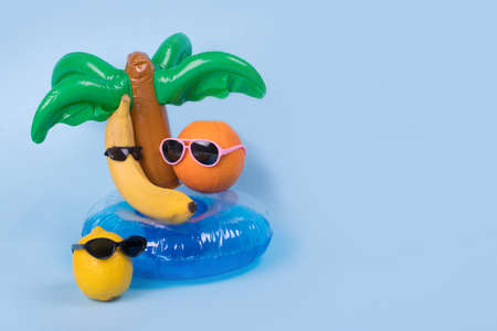 Creative fun idea with a banana, orange and lemon in sunglasses lying on a palm tree inflatable ring on a blue background. Minimal travel concept, summer stylish tropical fruit. Summertime color mood.Fashion, party and trend. Copy space.の写真素材