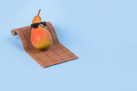 Minimal Fun idea made with a pear in sunglasses lying on a sunbed on a blue background. Trendy invitations with stylized fruit with space for text. Creative art, fashionable vacation, travel and fun concept.の写真素材