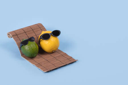 Creative fun idea with a lemon and lime in sunglasses lying on a sun bed on a blue background. Minimal travel and vacation concept, summer stylish tropical fruit. Summertime color mood. Fashion, holiday, party and summer trend. Copy space.の写真素材