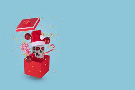 Gift box with human skull with Santa Claus hat, Christmas bauble balls and confetti on  bright blue background. Minimal New Year's seasonal concept. Creative Holiday greeting card.の写真素材