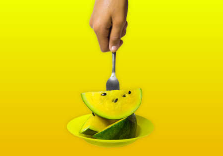 hand holding a fork stuck in a delicious and healthy yellow watermelon isolated on an orange yellow background. the benefits of watermelon help heart health and can nourish bodyの写真素材