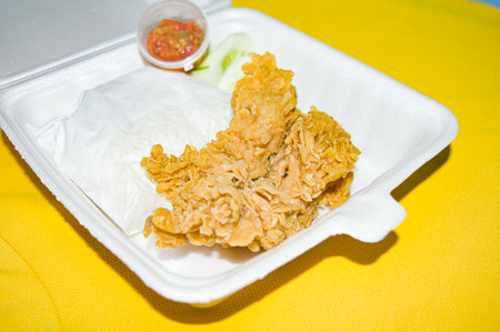close up, fried chicken with cucumber slices and raw chili sauce on a white styrofoam plate, with a yellow cloth background. Focus on fried chicken.の写真素材
