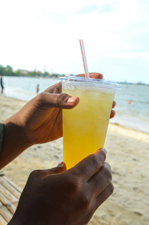 Hand holding iced lemonade, lime , orange juice in a plastic cup with a blurred beach background in...