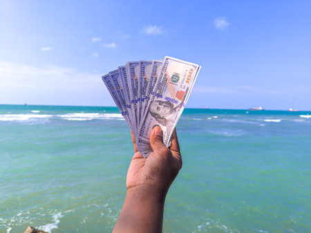Hand holding 100 dollar bill in hand, financial concept, vacation, success, salary. Summer sea water background. focus on dollar bill.の写真素材