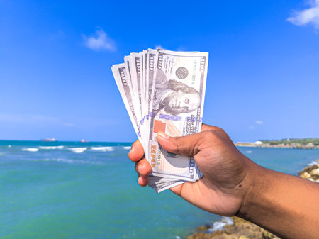Hand holding 100 dollar bill in hand, financial concept, vacation, tax, success, salary. Summer sea water background. focus on dollar bill.noisy, exposure, similar othersの写真素材