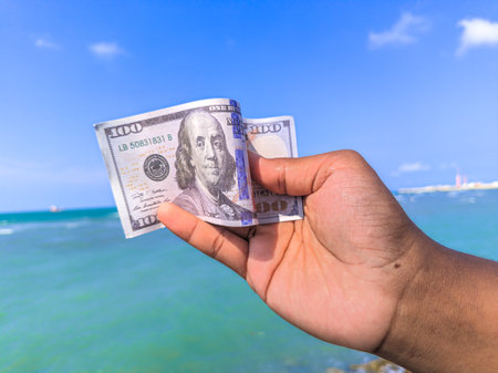 Hand holding 100 dollar bill in hand, financial concept, vacation, success, salary. Summer sea water background. focus on dollar bill.の写真素材