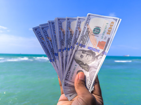 Hand holding 100 dollar bill in hand, financial concept, vacation, success, salary. Summer sea water background. focus on dollar bill.の写真素材