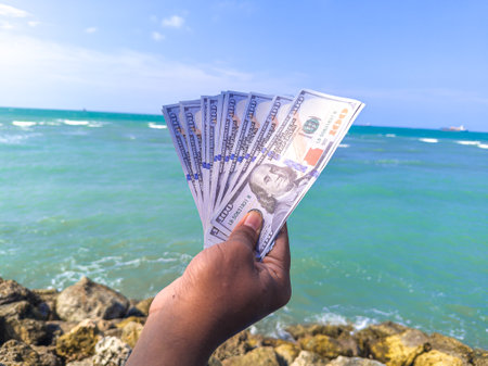 Hand holding 100 dollar bill in hand, financial concept, vacation, success, salary. Summer sea water background. focus on dollar bill.の写真素材