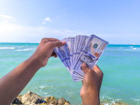 Hand holding 100 dollar bill in hand, financial concept, vacation, success, salary. Summer sea water background. focus on dollar bill.の写真素材