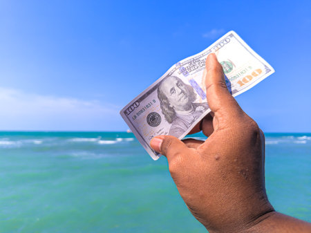 Hand holding 100 dollar bill in hand, financial concept, vacation, tax, success, salary. Summer sea water background. focus on dollar bill.noisy, exposure, similar othersの写真素材