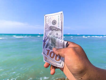 Hand holding 100 dollar bill in hand, financial concept, vacation, tax, success, salary. Summer sea water background. focus on dollar bill.noisy, exposure, similar othersの写真素材